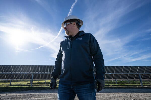 A man in a hard hat, goggles and a blue jacket under a blue sky, with a solar array in the background.