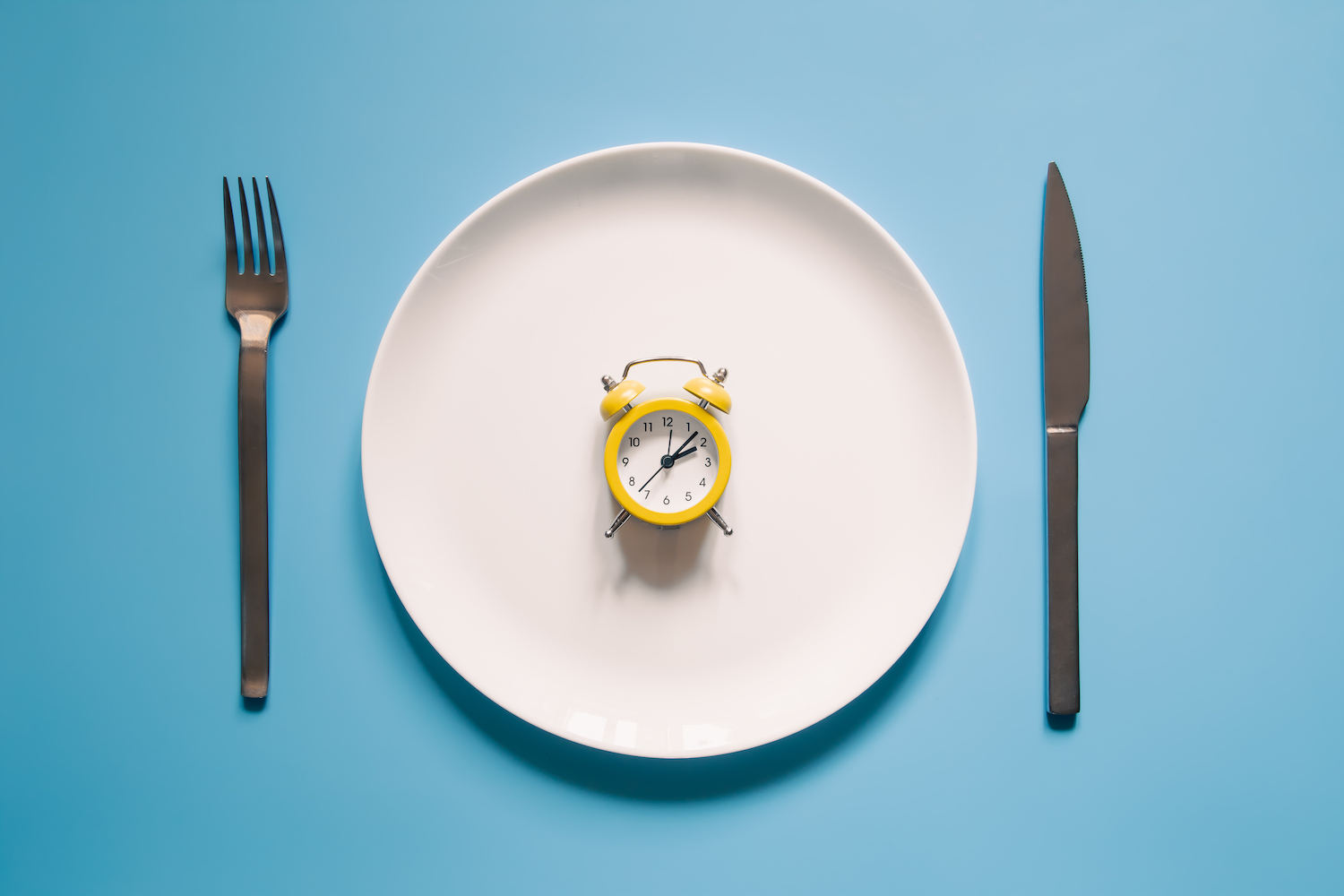 Alarm clock on a white plate with a knife and fork on blue background. Intermittent fasting, Ketogenic dieting, weight loss, meal plan, and healthy food concept.