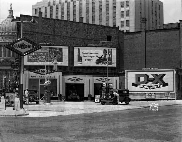 A black-and-white photograph of a Diamond gas station in Madison, Wis., in 1935. Two men are servicing cars and various billboards surround the station, one promoting Ethyl.