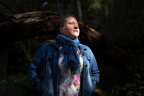 A woman in a jean jacket, blue patterned scarf and flowered shirt poses in front of a large log.