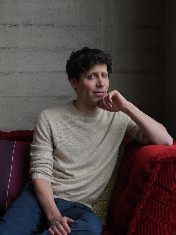 Sam Altman sits at the end of a red corduroy sofa, his left hand curled against his chin and his left elbow resting on the arm of the sofa.