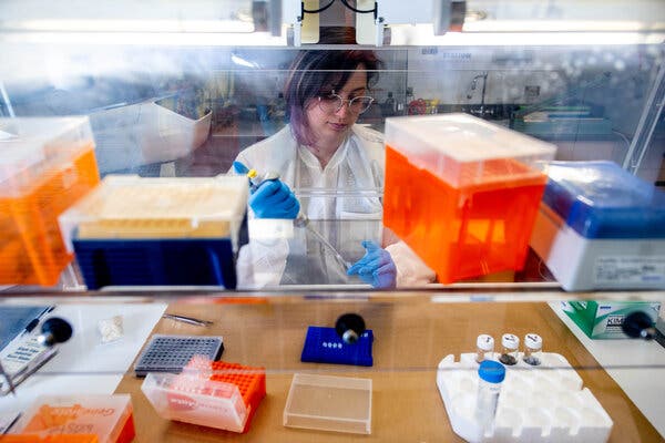 A woman in a white lab coat holds a metal tool. In front of her are various orange and blue plastic containers.