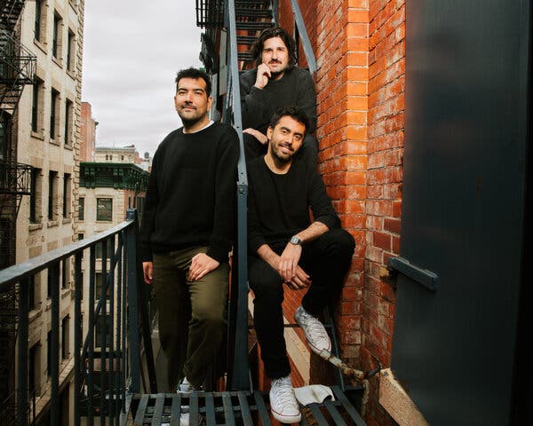 Three men in dark shirts pose on a fire escape on the side of a brick building.