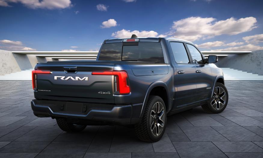 Back view of the upcoming Ram 1500 REV electric truck. It has a dark blue color and is sitting in a modern landscape with tiles and planks in the distance (with a blue sky behind it).