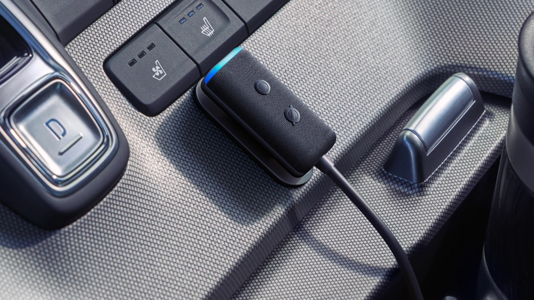 Marketing photo of the second-gen Amazon Echo Auto. The thin gray speaker has a blue light glowing at its top and two buttons on its fabric-covered body. It's mounted on a car's center console.