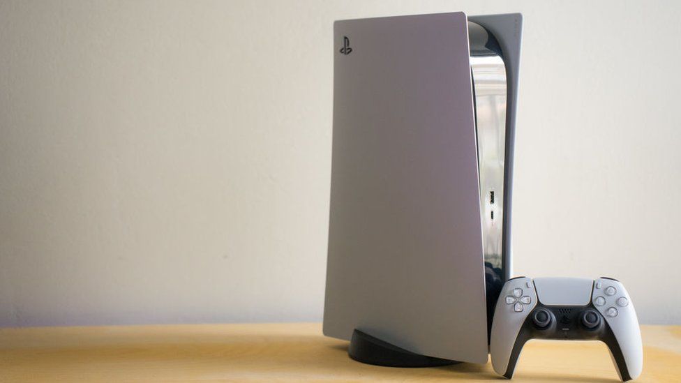 A PS5 console, stood vertically on a plain wooden table top in front of a white wall. A Dualsense controller is propped up against the console