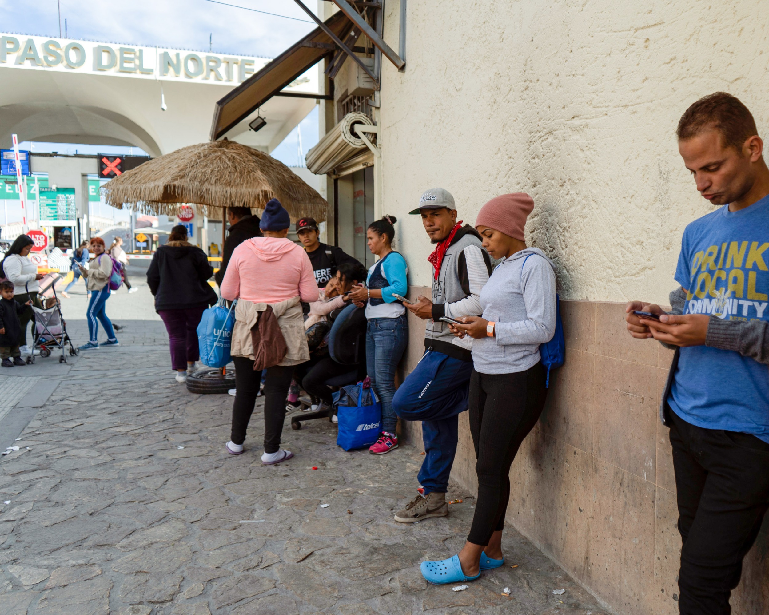 Keisy P leans against the wall of a building with others as they check their phones.