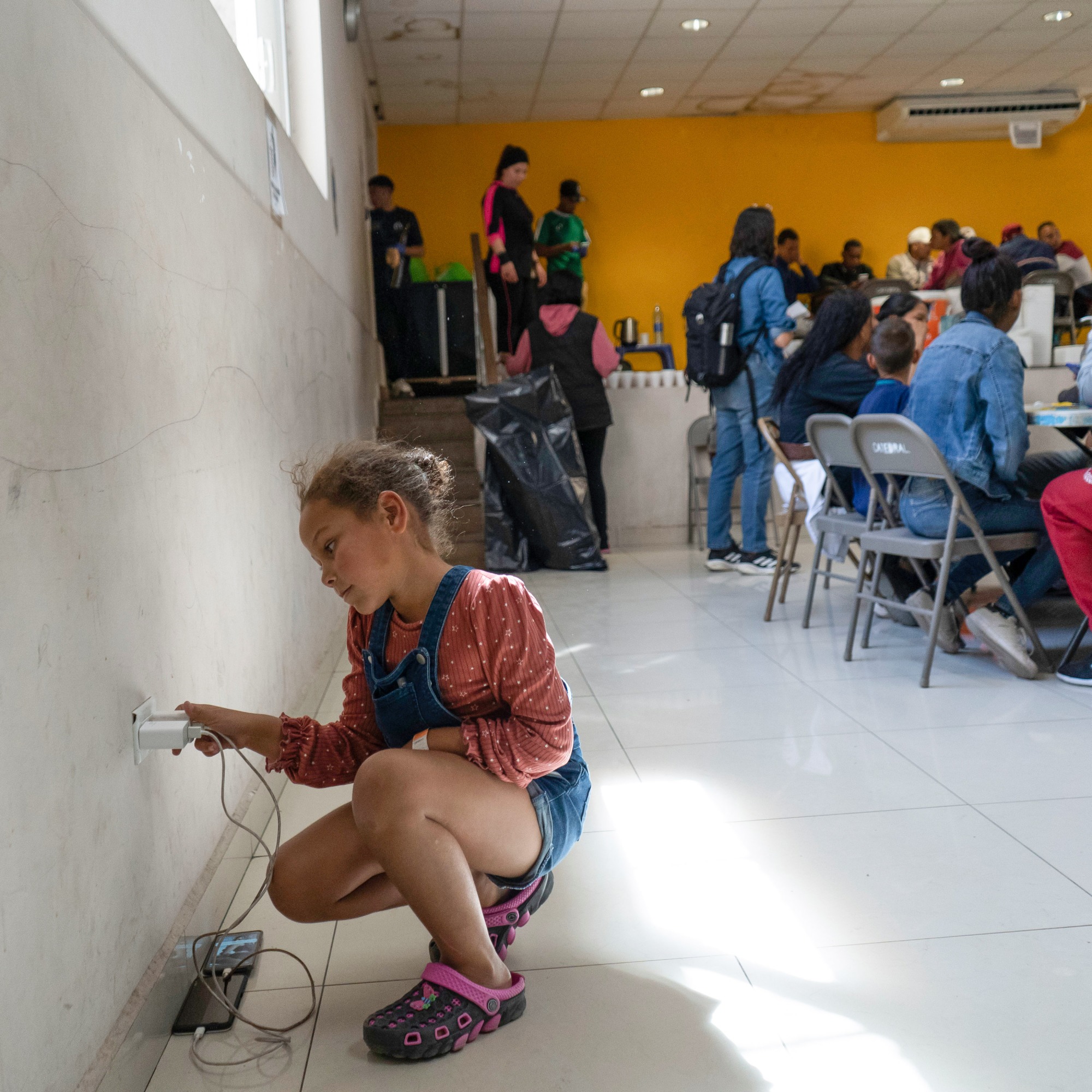 Arantza Plaza kneels on the floor next to phones plugged into the wall to charge while adults meet at a table in the room behind her.