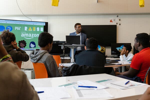 A man wearing a blazer stands in the middle of a classroom talking with high school students. A screen behind him says “Amazon.” 