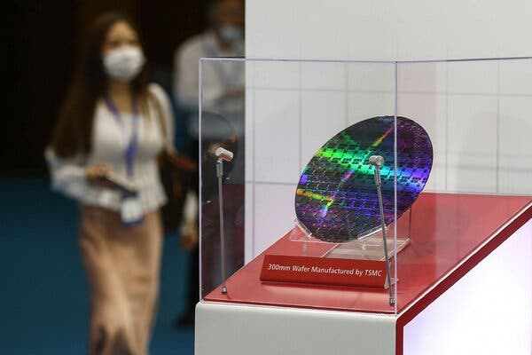 A silicon wafer is displayed in a clear glass case with a label that reads “300mm Wafer Manufactured by TSMC.” A woman and a man are in the background, both wearing lanyards and face masks.