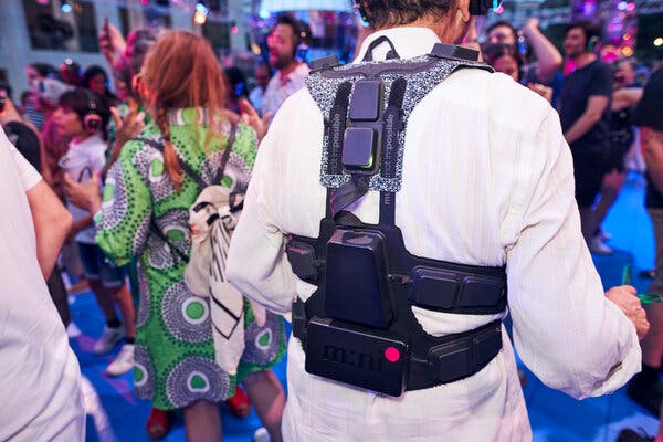 A person wearing a white shirt stands in a crowd of people dancing at a party. The person is wearing a black device called a haptic suit that wraps around the shoulders and the waist like a backpack. 