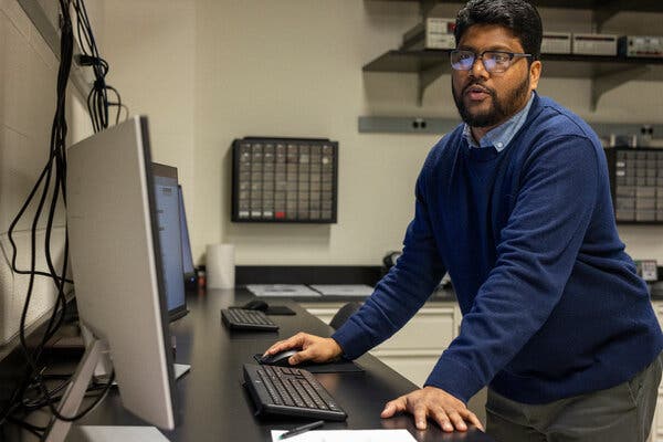 Ranga P. Dias wears a blue sweater over a blue collared shirt and stands looking at a computer on a black counter in a lab.