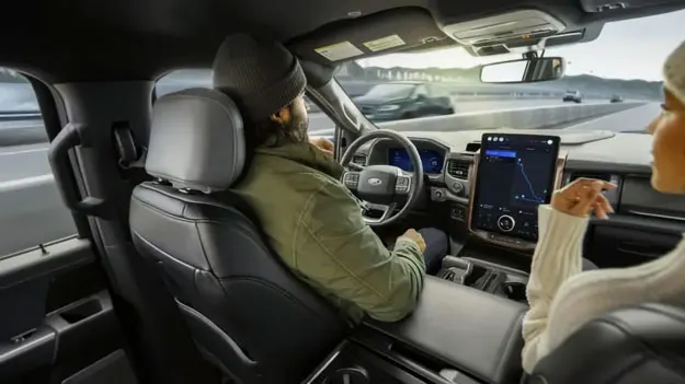 Promotional image of one of Ford's new tank-like vehicles with a massive screen in the middle and two people in the front seats, a hefty, bearded figure behind the wheel and a white-clad person on the right of the shot who is barely in focus.