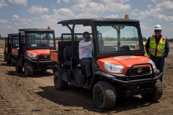 Brad Chambers sits in one of four small orange vehicles in a dirt field. Standing next to him is a worker in a construction hat and neon yellow vest. 