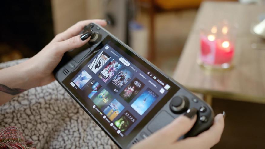 Photo of the Steam Deck portable console in hand. Two hands hold the device, which shows a game library. A blurred coffee table sits in the background.