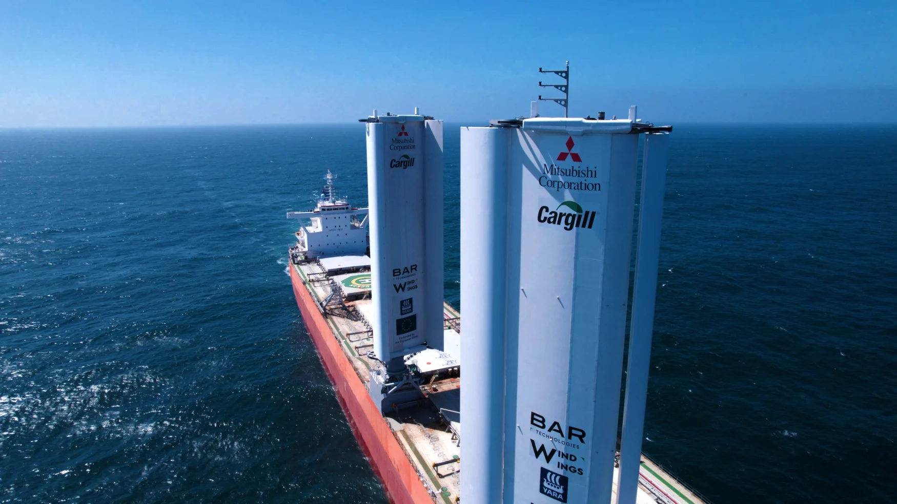 A cargo ship fitted with two enormous, rigid sails. Bird's eye view looking down from near the top of one of the sails (both are visible with most of the ship's deck).