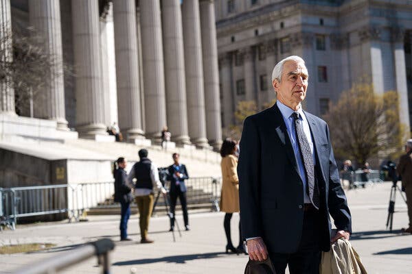 Sam Waterston, wearing a dark suit and tie, stands on the sidewalk in front of a courthouse.