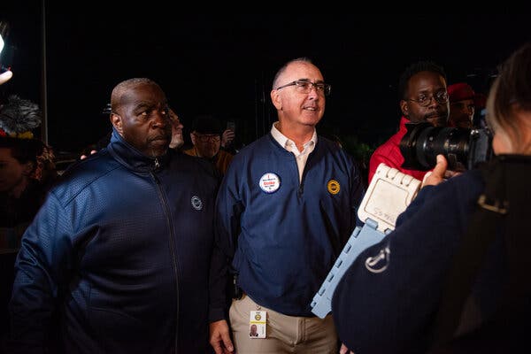 Shawn Fain stands with union workers while someone in the foreground takes their photograph with a camera.