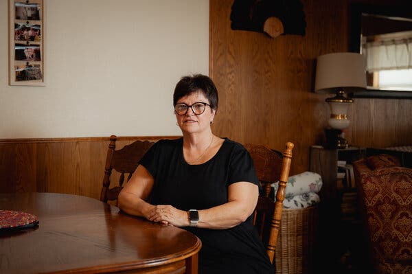 A woman in a black short-sleeved T-shirt sits at a dining room table in a wood-paneled home.