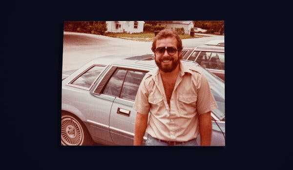 A sepia-toned photograph of Hank Asher in a tan, short-sleeved button down shirt standing in front of a car.