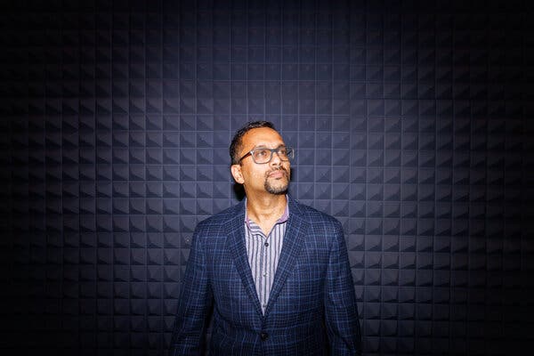 Vijay Balasubramaniyan, in a blue plaid jacket, stands in front of a dark patterned wall looking up at an angle.