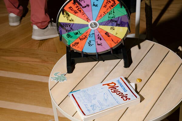 A paperback book titled “Thinking Physics” lies on a small round table. A disc that can be spun, divided into colorful sections, stands behind it. Between them is a sticker of a green, octopus-like creature with many eyes. 