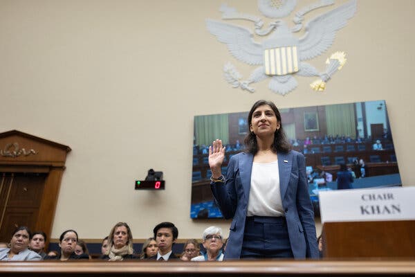 Lina Khan, wearing a blue suit and a white top, stands in court with her arm raised in the air. Behind her are seated audience members.