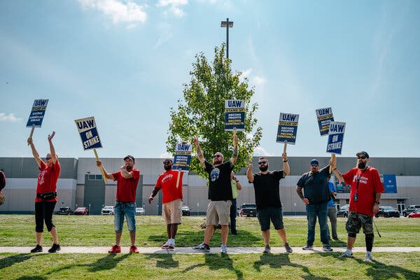 People standing near a white and gray building and holding signs that say “UAW on Strike” and “UAW Stand Up.”