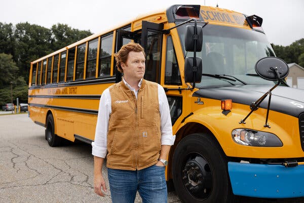 Duncan McIntyre wearing a white shirt, jeans and a brown vest with “Highland” written on it. He is standing in front of a school bus.