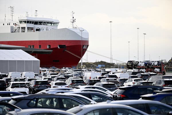 A large area is filled with cars with a tall red and white ship in the background.