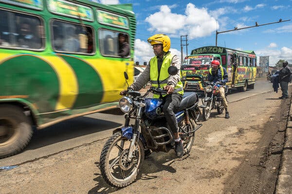 A man on a motorcycle wearing a green protective vest is in the foreground. Next to him is a bus and behind him is a motorcyclist who has stopped and is looking down.