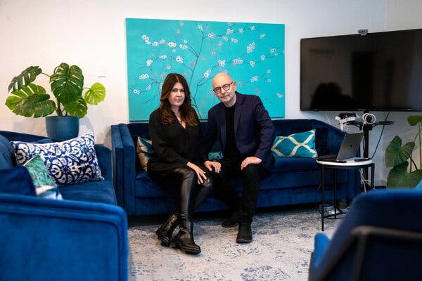 A woman with long dark brown hair, dressed in black, sits on a blue couch in an office next to a man with close-cropped hair and glasses.