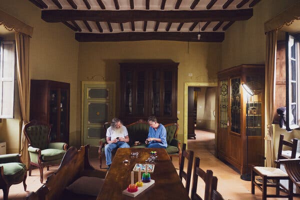 A wide view of the Millers sitting on an antique couch in a room with large, antique bureaus holding various puzzles and a long dining room table scattered with other puzzle specimen. 