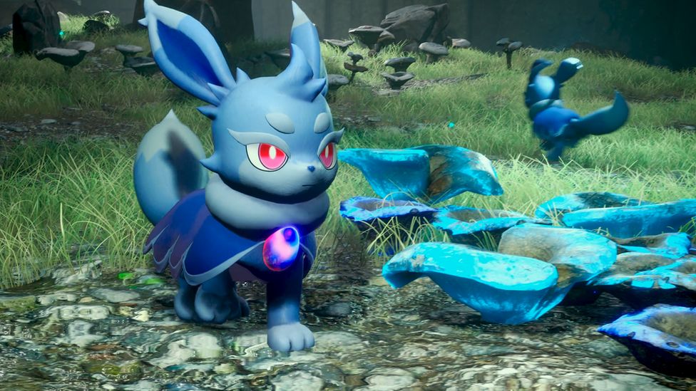 A cute, blue, bunny like creature with stern expression and ruby red pupils in its large eyes stands in a field of grass. It's next to some flowers that glow blue, suggesting some sort of magical property. The rabbit creature also wears a dark blue cloak, fastened below the next with a gem with swirling pink and blue glow.