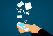 An image depicting ecommerce. A pair of hands hold a mobile phone and shopping icons like a dollar sign and a trolley float up from the screen